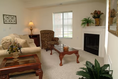 A cozy living room with a beige sofa, a patterned armchair, a wooden coffee table, and a side table with a lamp. There is a fireplace with decorative plants on the mantel and a window letting in natural light.