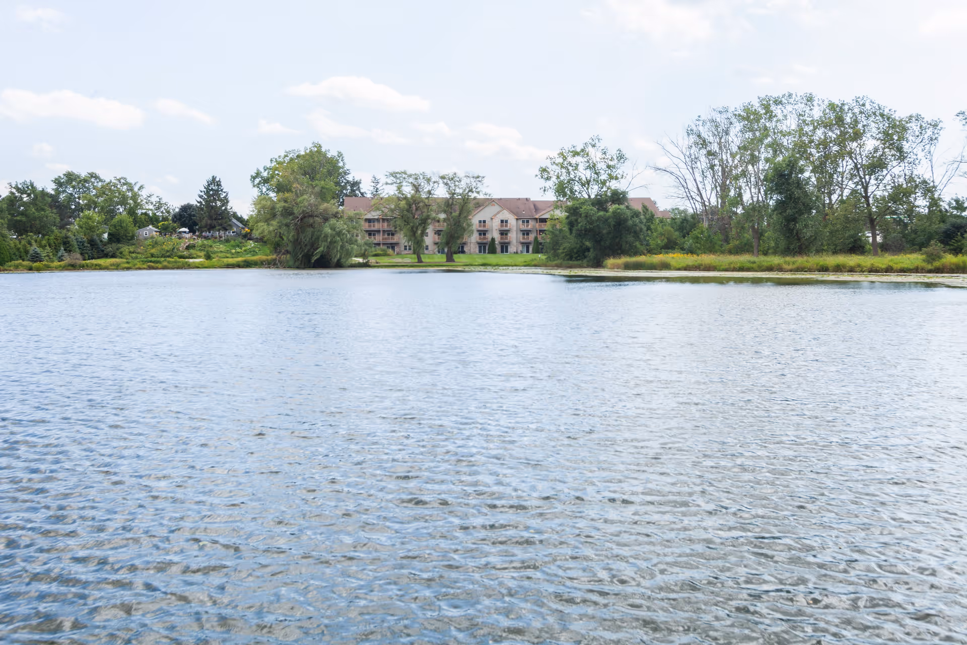 A large body of water with gentle ripples in the foreground, surrounded by green trees and vegetation. In the background, there is a multi-story building partially obscured by trees under a partly cloudy sky.