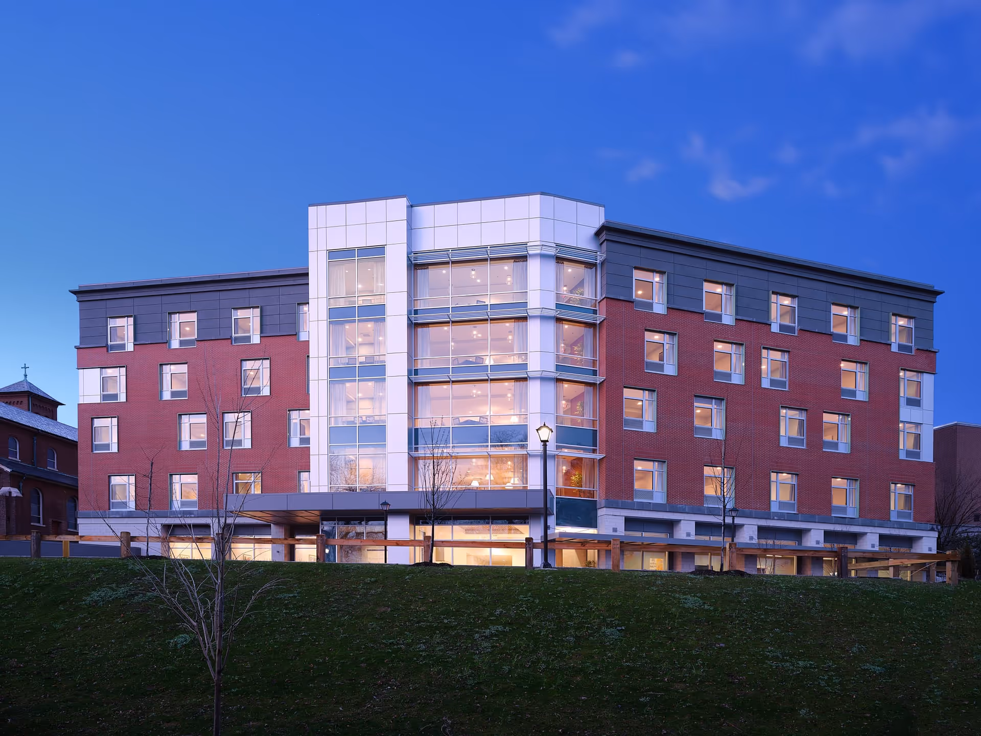 Exterior view of a modern multi-story senior living facility building named The Wartburg Home during twilight, with large windows and a well-lit interior, set against a clear blue sky and a grassy foreground.