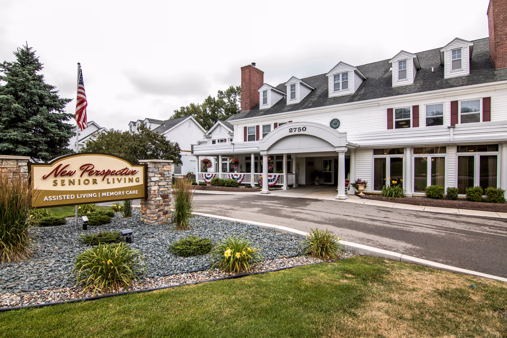 Exterior view of New Perspective Senior Living facility in Roseville, showing a white multi-story building with a covered entrance and the address number 2750. There is a landscaped area with plants and a sign that reads 'New Perspective Senior Living Assisted Living | Memory Care' near the driveway. An American flag is visible near the sign.