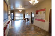A spacious hallway in a senior living facility with tiled flooring and beige walls. There are chandeliers hanging from the ceiling, a bulletin board with notices on the right wall, and a table with chairs near the far end by large windows letting in natural light.