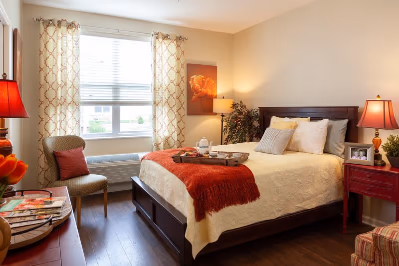 A cozy bedroom with a large window covered by patterned curtains letting in natural light. The room features a wooden bed with cream and gray pillows, a red throw blanket, and a tray with a teapot and cups. There are two red lamps on either side of the bed, a chair with a red cushion near the window, a small red nightstand with a framed photo and a plant, and a wooden floor.