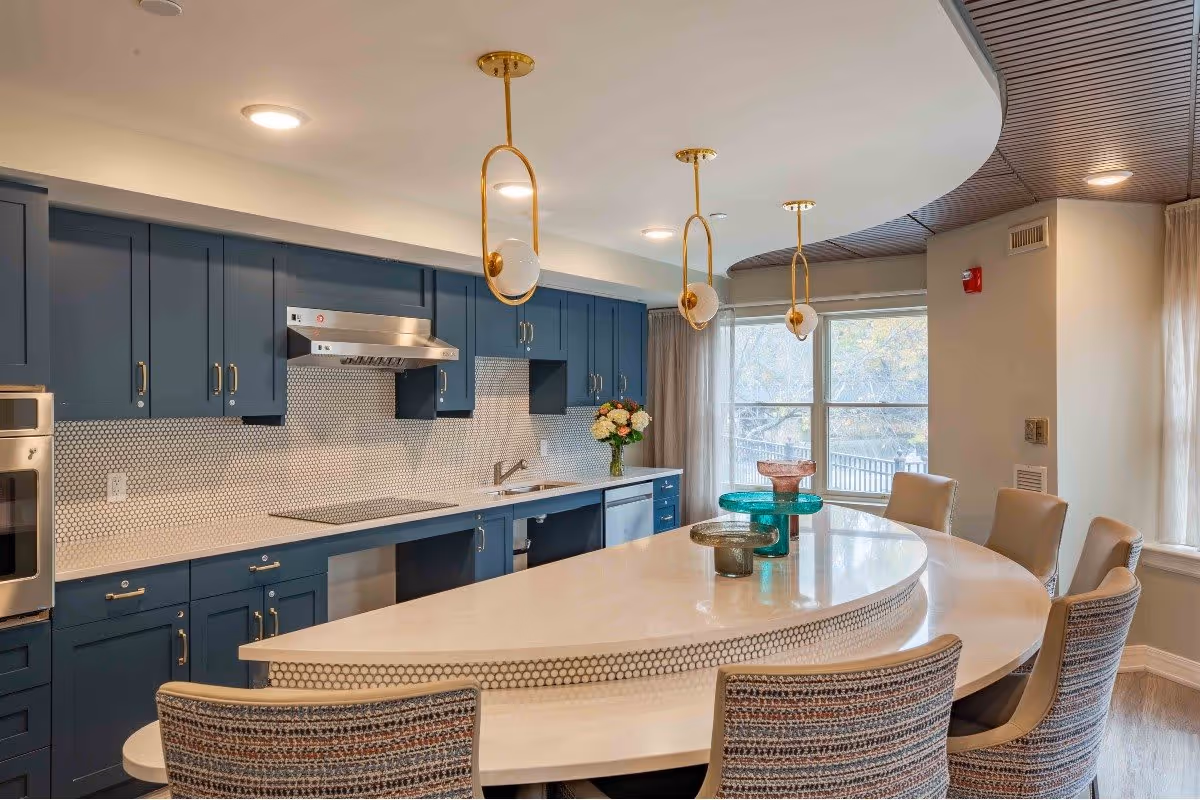Modern kitchen with blue cabinets, a white countertop island with seating, three pendant lights hanging above, and a window with curtains letting in natural light.