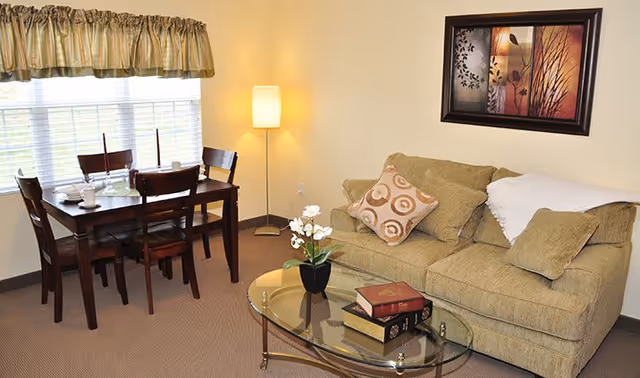 A cozy living room and dining area with a beige couch adorned with patterned and plain pillows, a glass coffee table with books and a small potted plant, a wooden dining table set with four chairs near a window with striped valance curtains, and a floor lamp in the corner. A framed artwork hangs on the wall above the couch.