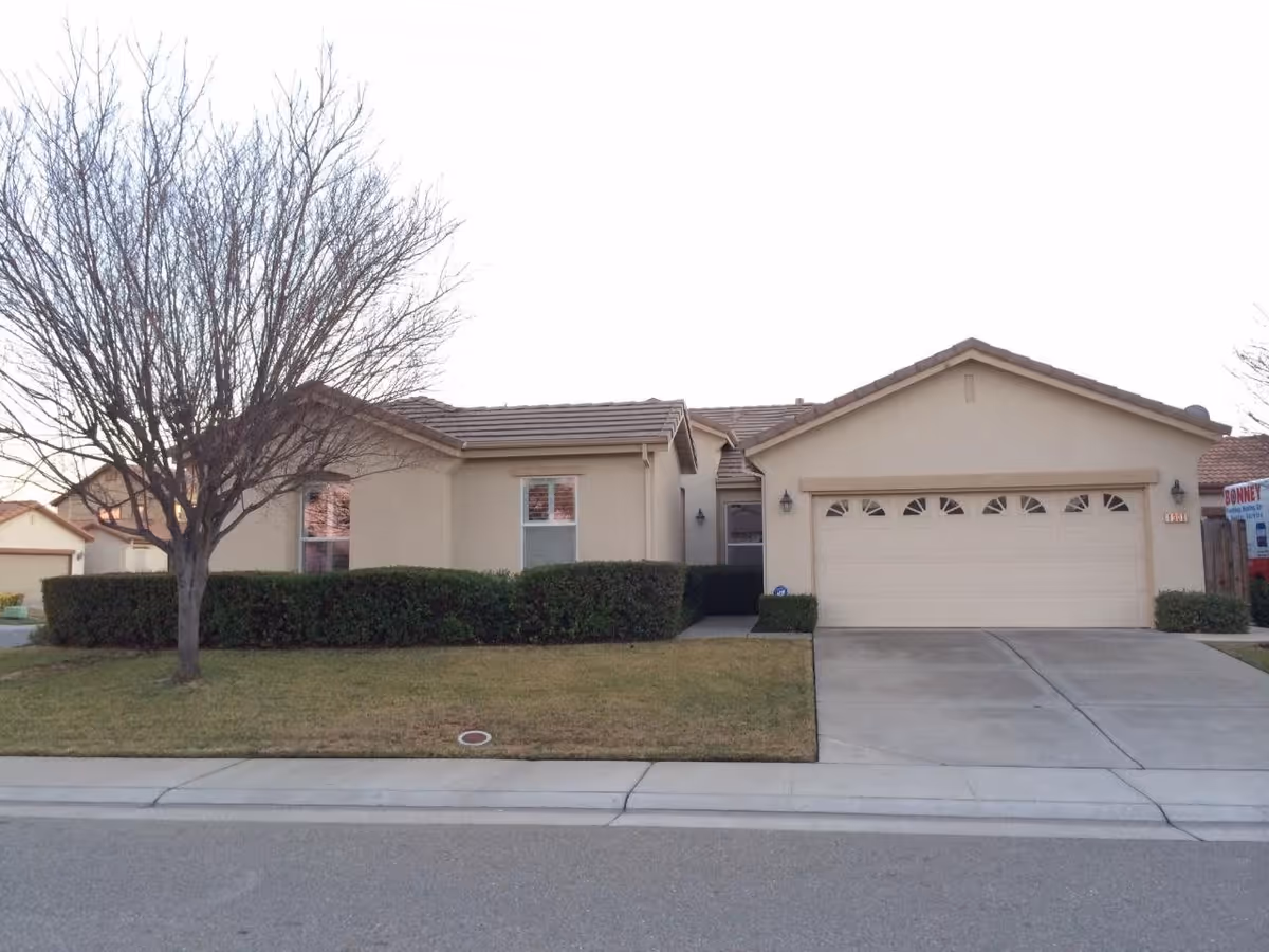 Single-story suburban house with an attached two-car garage, driveway, front lawn, hedges, and a leafless tree.