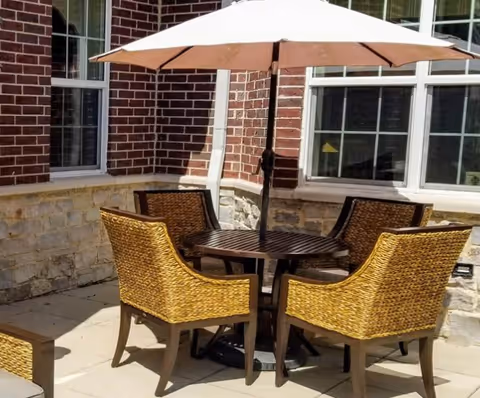 Outdoor patio area with a round wooden table and four wicker chairs arranged around it. A large beige umbrella is positioned in the center of the table providing shade. The patio is adjacent to a brick and stone building with windows.
