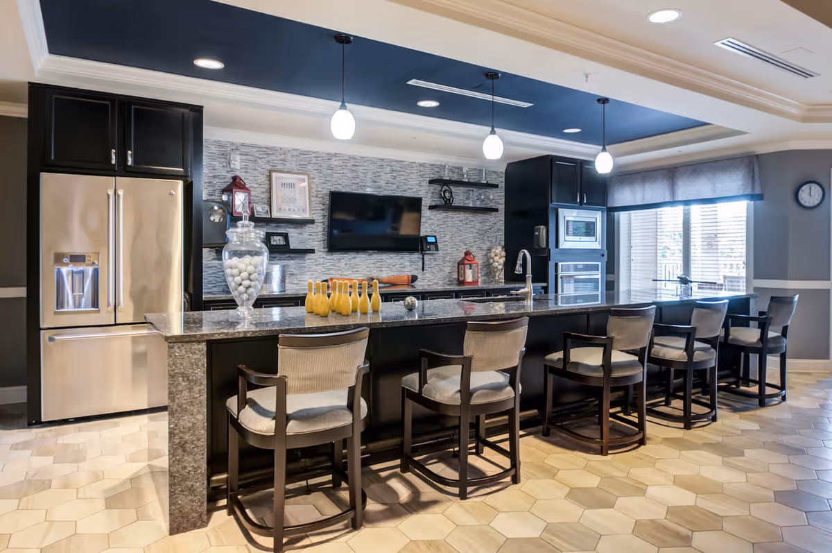Modern kitchen area with a long granite countertop island featuring six cushioned bar stools. The kitchen has dark cabinetry, stainless steel refrigerator, built-in microwave and oven, pendant lights hanging from a dark blue ceiling section, and a wall-mounted TV. Decorative items including jars filled with white balls and small bowling pins are placed on the countertop. Large windows with blinds allow natural light into the space.