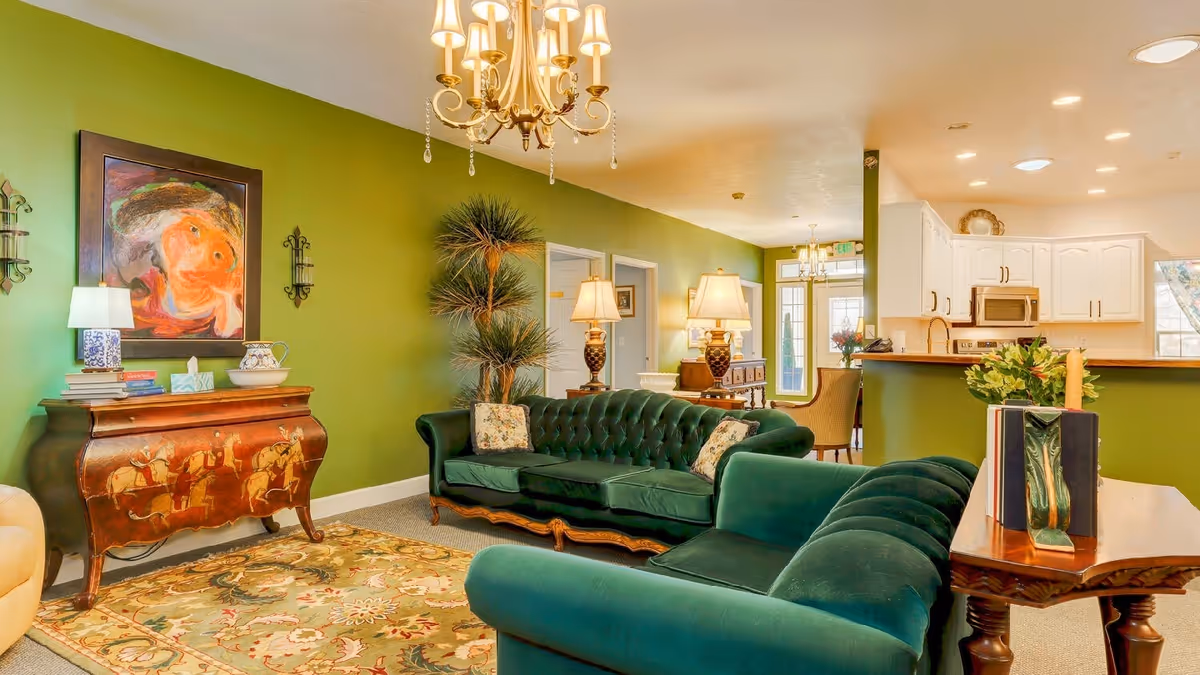 A cozy living room with green walls and two dark green velvet sofas facing each other. There is a wooden side table with books and a decorative vase, a vintage wooden chest with a painting above it, and a tall plant in the corner. The room is well-lit with table lamps and a chandelier. In the background, a white kitchen with cabinets and a countertop is visible.