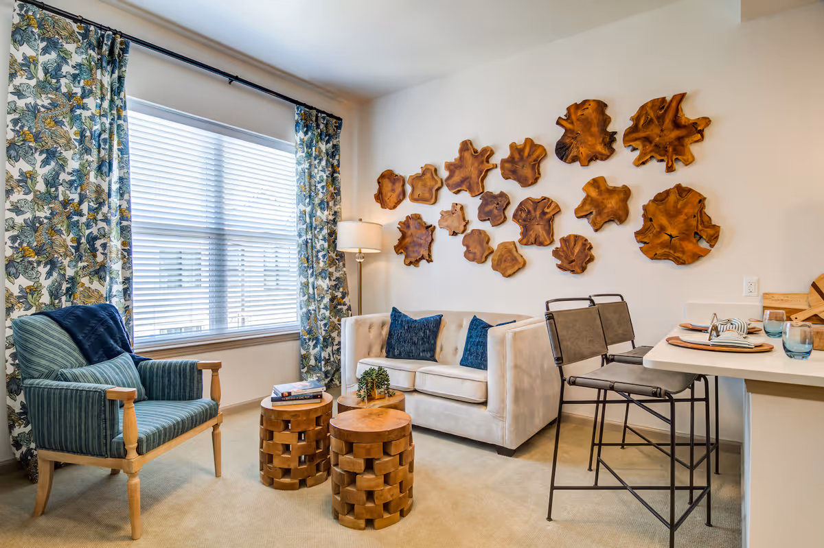 A cozy living room with a beige sofa adorned with blue pillows, a blue striped armchair, and two wooden round coffee tables. The wall behind the sofa features decorative wooden art pieces resembling tree slices. A large window with floral patterned curtains allows natural light to fill the room. To the right, there is a small dining area with two high chairs and a table set with plates and glasses.