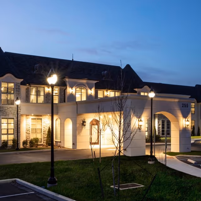 Exterior view of The Remington of Yardley senior living facility at dusk, showing a well-lit entrance with arched doorway, stone and stucco facade, street lamps, and a small landscaped area with young trees and grass.