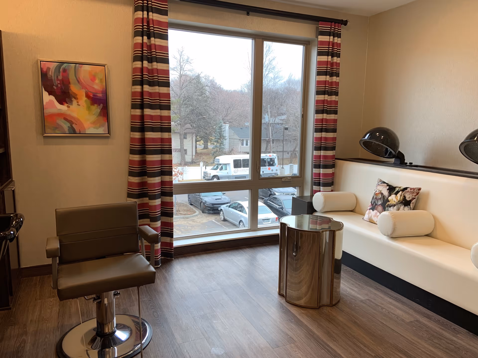A cozy interior room with a large window showing a view of parked cars and trees outside. Inside, there is a brown salon chair, a white cushioned bench with floral and cylindrical pillows, two black hair dryer hoods mounted on the wall, a small round metallic table, and a colorful abstract painting on the wall. The room has wooden flooring and striped curtains framing the window.