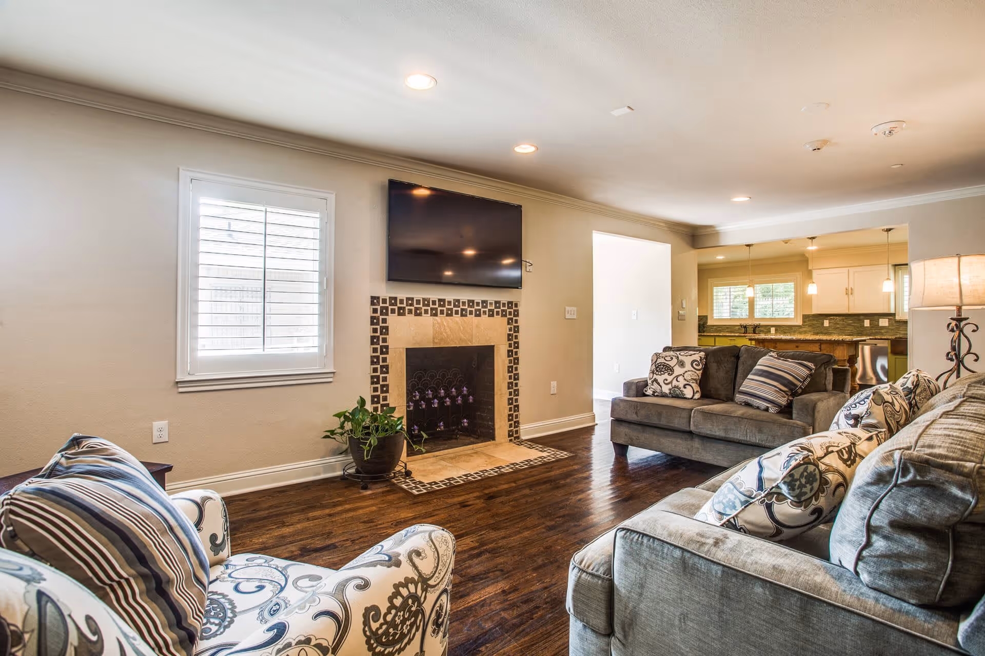 Bright living room with hardwood floors, patterned armchairs and sofas, a tiled fireplace with wall-mounted TV, and an open view into the kitchen.