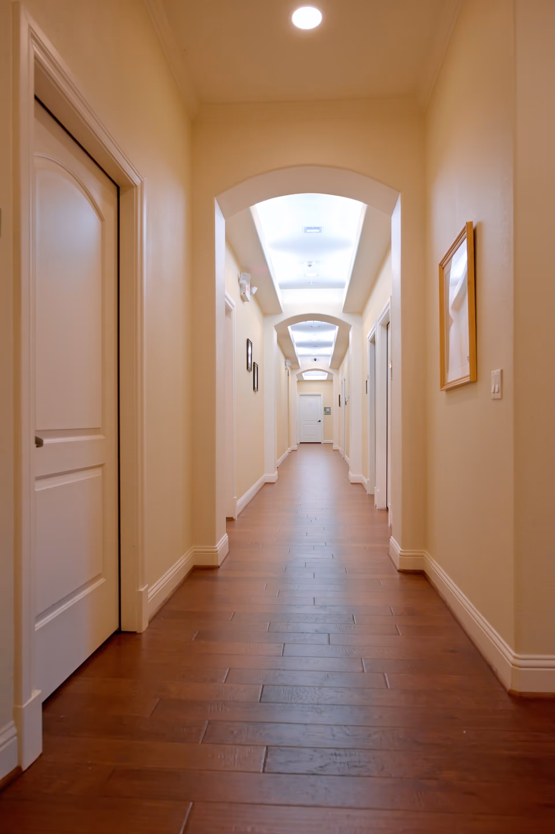 A long, well-lit hallway with wooden flooring and cream-colored walls. The hallway features several closed white doors on both sides and framed pictures hanging on the walls. The ceiling has recessed lighting and decorative arches.