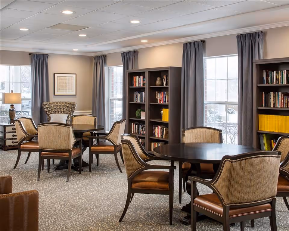 A cozy senior living common area with round wooden tables surrounded by cushioned chairs. The room features large windows with gray curtains, bookshelves filled with books, a patterned armchair, a side table with a lamp, and framed artwork on the wall. The carpet is patterned and the ceiling has recessed lighting.