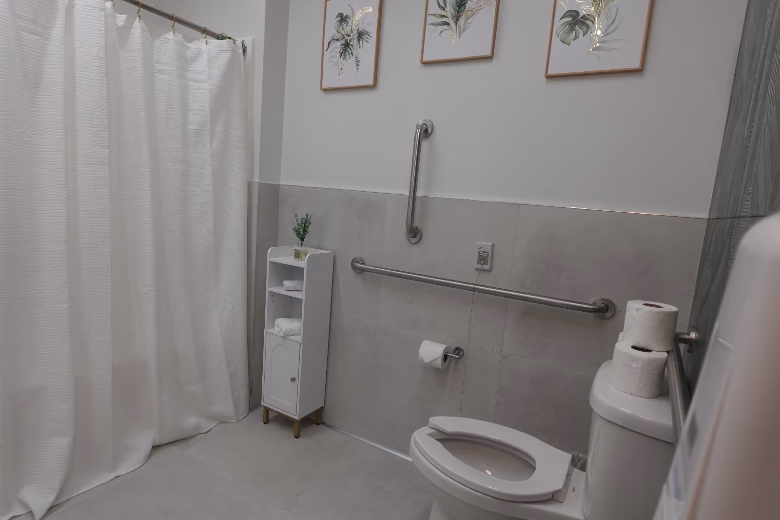 A clean and accessible bathroom with a white shower curtain, a white toilet with two rolls of toilet paper on top, a toilet paper holder with a roll, and grab bars on the tiled wall. There is a small white cabinet with shelves holding towels and a small plant. Three framed botanical prints hang on the wall above the grab bars.