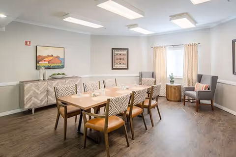 A bright dining room featuring a rectangular wooden table with six chairs, a sideboard, framed artwork, and a window with curtains.