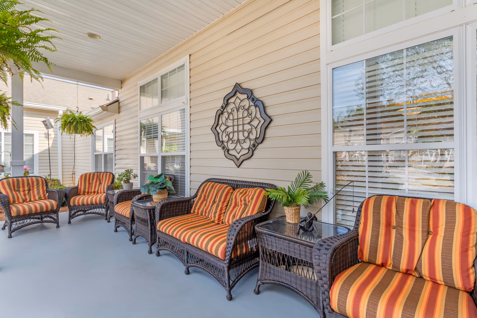 Covered outdoor patio area with wicker furniture including two armchairs, a loveseat, and two side tables. The furniture has cushions with orange, red, and brown stripes. There are potted plants on the tables and hanging ferns from the ceiling. The patio is adjacent to a beige siding building with large windows and a decorative metal wall hanging.