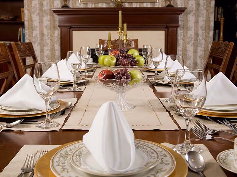 A formal dining table set with white folded napkins, wine glasses, plates, and silverware. The table has a beige runner with two glass bowls filled with green apples, red apples, and grapes as centerpieces. In the background, there is a wooden fireplace mantel with decorative candlesticks.