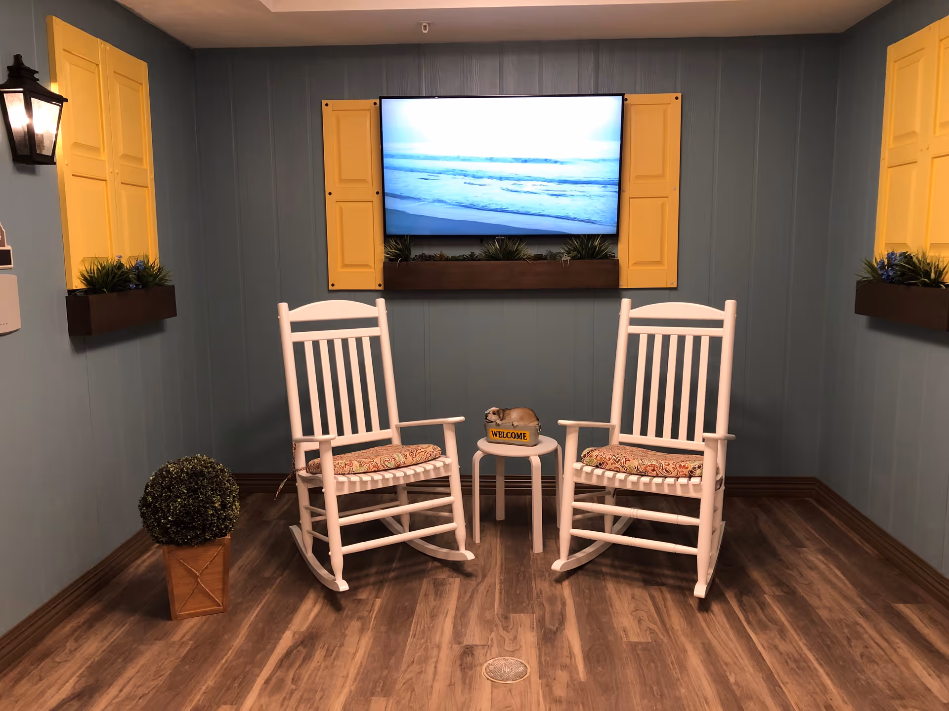 A cozy sitting area with two white wooden rocking chairs with patterned cushions, a small white side table between them holding a small dog figurine with a welcome sign. The walls are painted blue with yellow window shutters and planter boxes with green plants. A flat-screen TV mounted on the wall displays an ocean scene. The floor is wood with a warm tone.