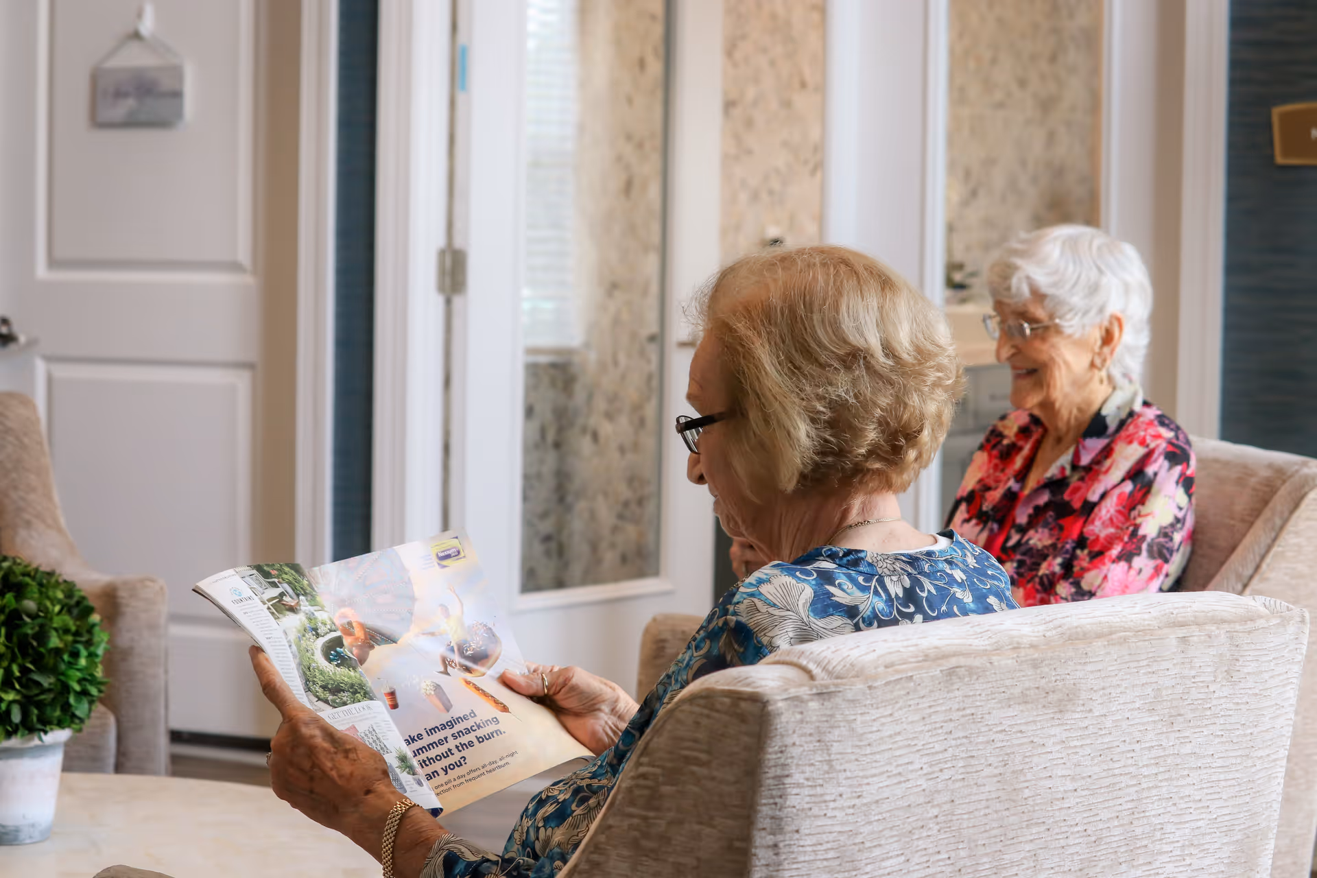 Two elderly women sitting in a cozy living room area. One woman is reading a magazine while the other is smiling and looking in her direction. The room has light-colored furniture, a small green plant on a table, and a door with glass panels in the background.
