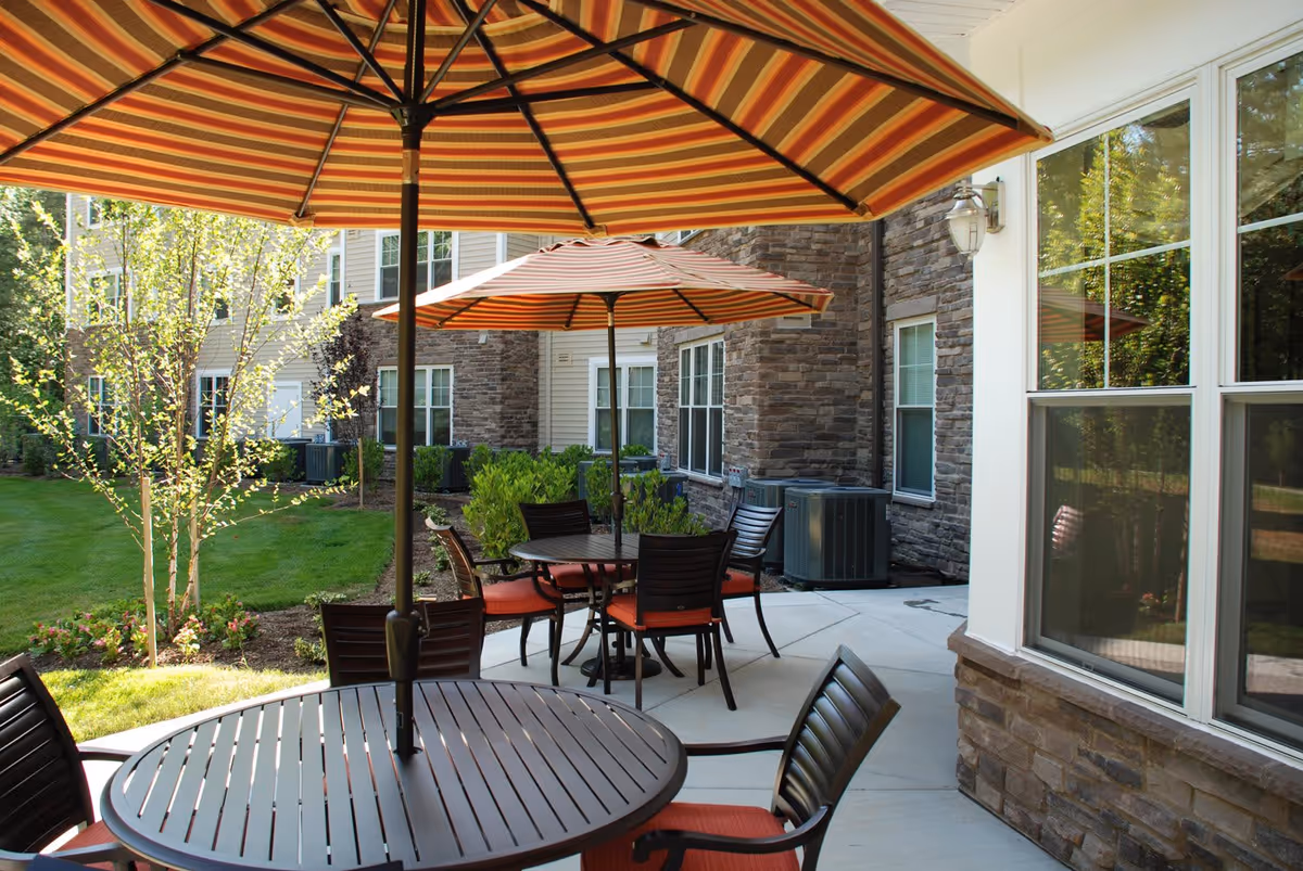 Outdoor patio area with round tables and chairs featuring orange cushions, shaded by large striped umbrellas. The patio is adjacent to a building with stone and siding exterior walls, and there is a well-maintained lawn and small trees in the background.