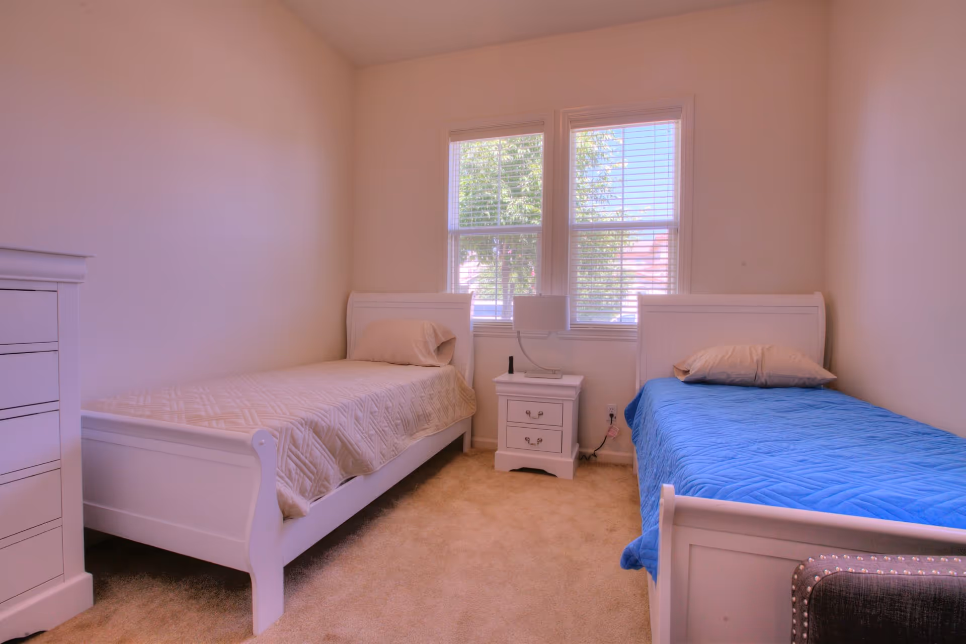 A bedroom with two single beds, one with a beige quilt and pillow and the other with a blue quilt and beige pillow. Between the beds is a white nightstand with a lamp and a remote control. There is a window with blinds behind the nightstand, and a white dresser is visible on the left side of the room. The carpet is beige and the walls are light-colored.