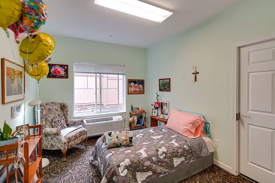 A cozy bedroom in a memory care facility with a single bed covered in a gray blanket featuring white dog patterns and a pink pillow. There is a floral armchair near a window with blinds, a small wooden side table with framed photos and flowers, and several colorful balloons with smiley faces attached to a floor lamp. The walls are painted light green and decorated with framed pictures and a cross. The floor has a patterned carpet.