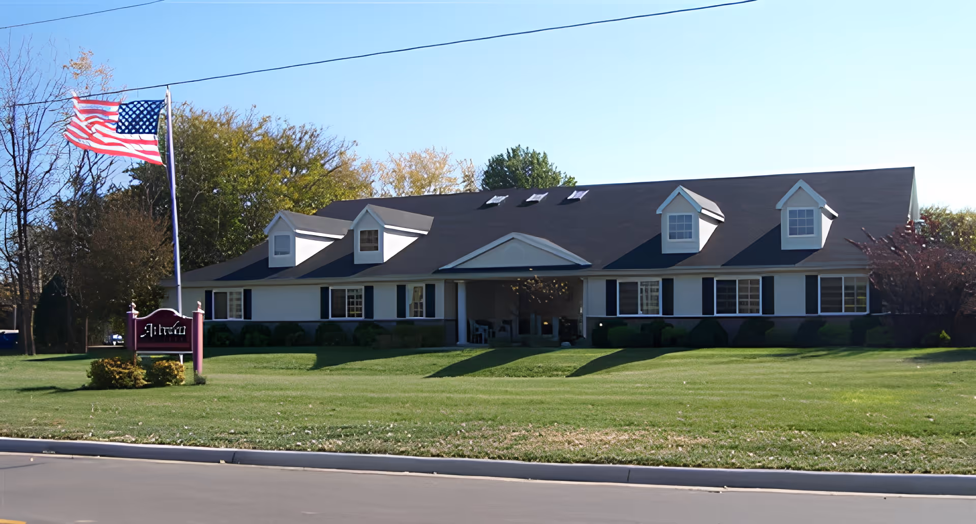 Exterior view of a single-story building with a dark roof and white walls, featuring several dormer windows. In front of the building is a well-maintained lawn, an American flag on a flagpole, and a sign that reads 'Avonlea'. Trees with autumn foliage are visible in the background under a clear blue sky.