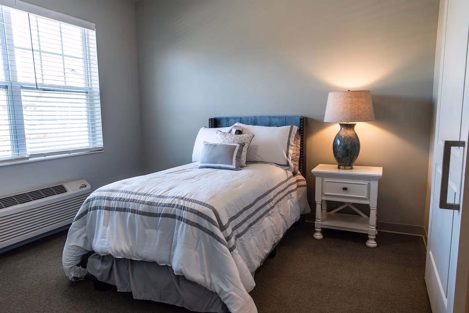 A neatly made single bed with white and gray striped bedding and decorative pillows in a small bedroom. Next to the bed is a white nightstand with a blue ceramic lamp turned on. A window with white blinds is on the left wall, and a white wardrobe is on the right.