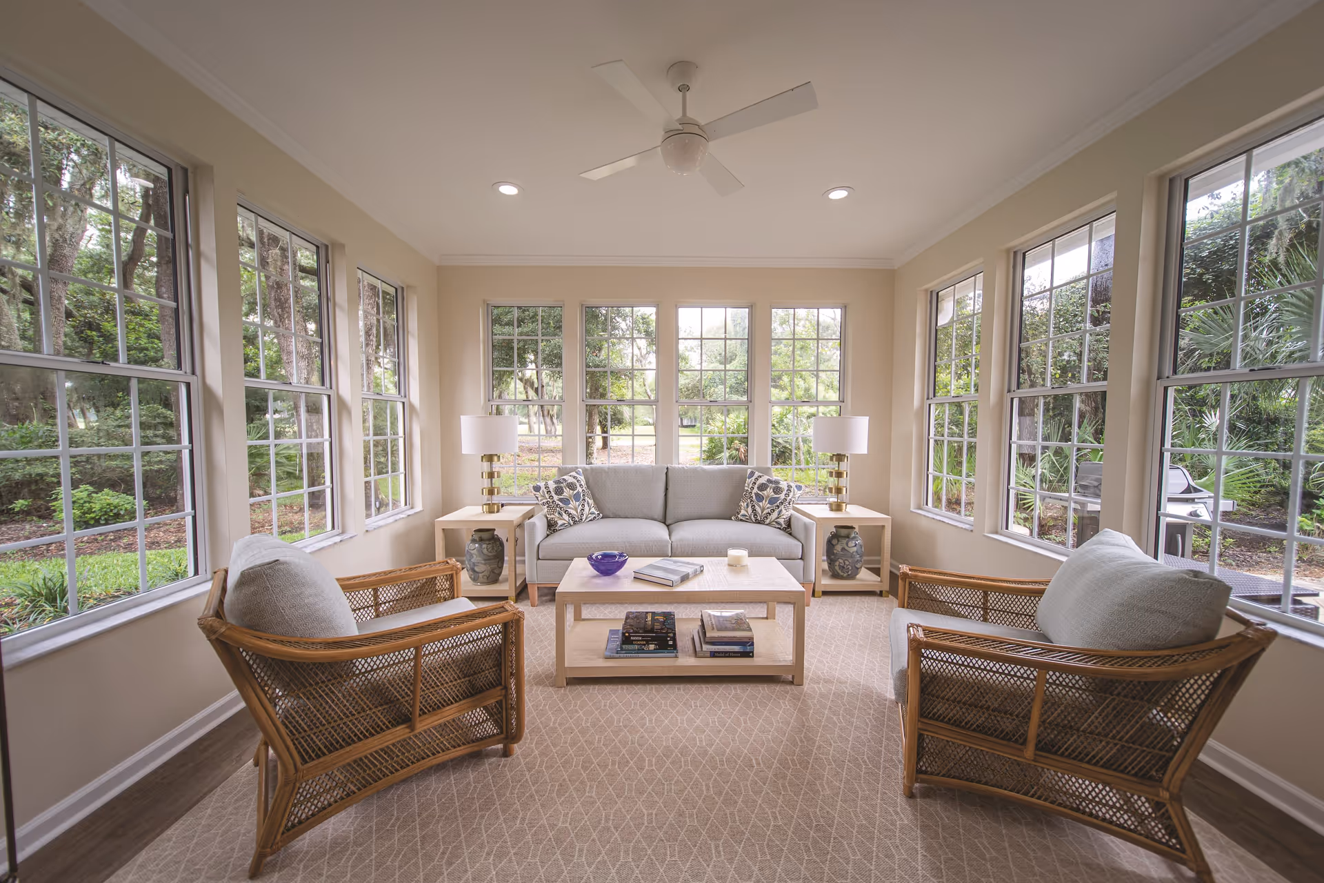 A bright and cozy living room with large windows on three sides showing a green outdoor view. The room features a light gray sofa with patterned cushions, two wicker armchairs with cushions, a light wood coffee table with books and decorative items, two matching side tables with lamps, and a ceiling fan above.
