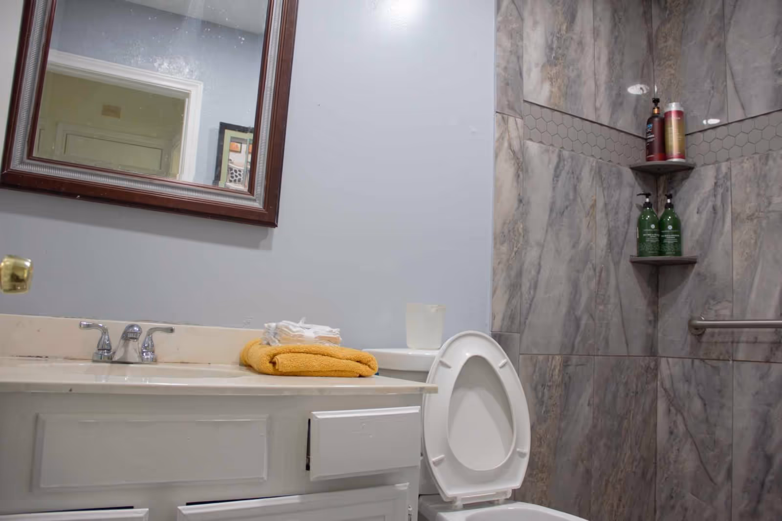 Bathroom with a white toilet with the lid open, a white vanity with a sink, a mirror above the sink, and a shower area with gray marble tiles and corner shelves holding bottles of shampoo and soap.