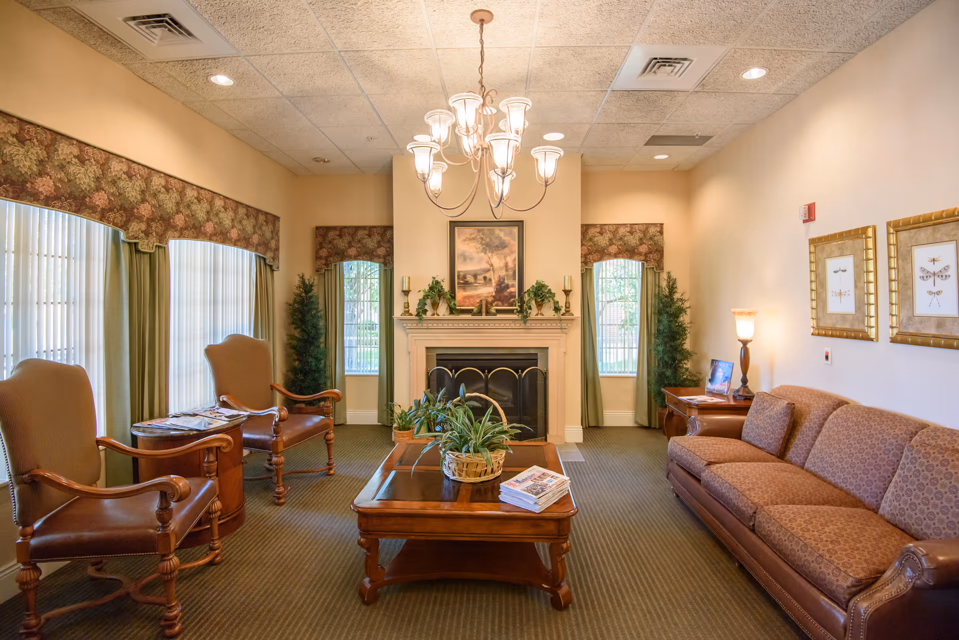A cozy living room area with a brown patterned sofa, two wooden armchairs with leather seats, a wooden coffee table with plants and magazines, a fireplace with decorative plants and a framed picture above it, floral valances on the windows, and warm lighting from a chandelier and a table lamp.