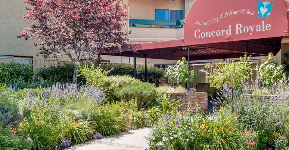 Outdoor garden area with various green plants and flowers in front of a building. A red canopy with the text 'Assisted Living With Heart & Soul Concord Royale' is visible, providing shade to a seating area underneath.