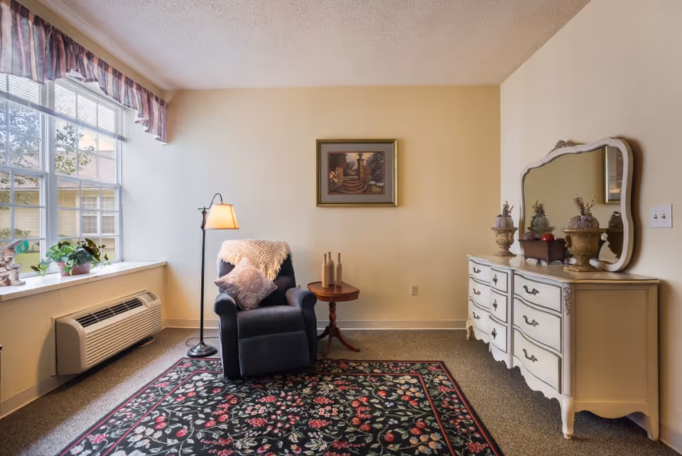 A cozy sitting area in a room with a large window on the left letting in natural light. The room features a dark upholstered armchair with a throw blanket and pillow, a small wooden side table with decorative bottles, a floor lamp, a floral patterned area rug, and a white dresser with an ornate mirror and decorative items on top. The walls are painted light beige and there is a framed picture hanging above the chair.