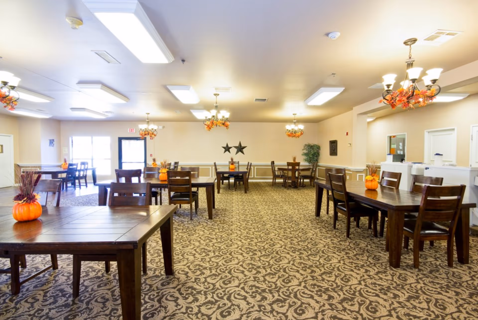 A spacious dining room with multiple wooden tables and chairs arranged neatly. Each table has a small pumpkin centerpiece with autumn-themed decorations. The room has patterned carpet flooring, beige walls with white trim, and several chandeliers with warm lighting. There are two black star decorations on the far wall and a potted plant in the corner. Large windows and a door provide natural light from one side.
