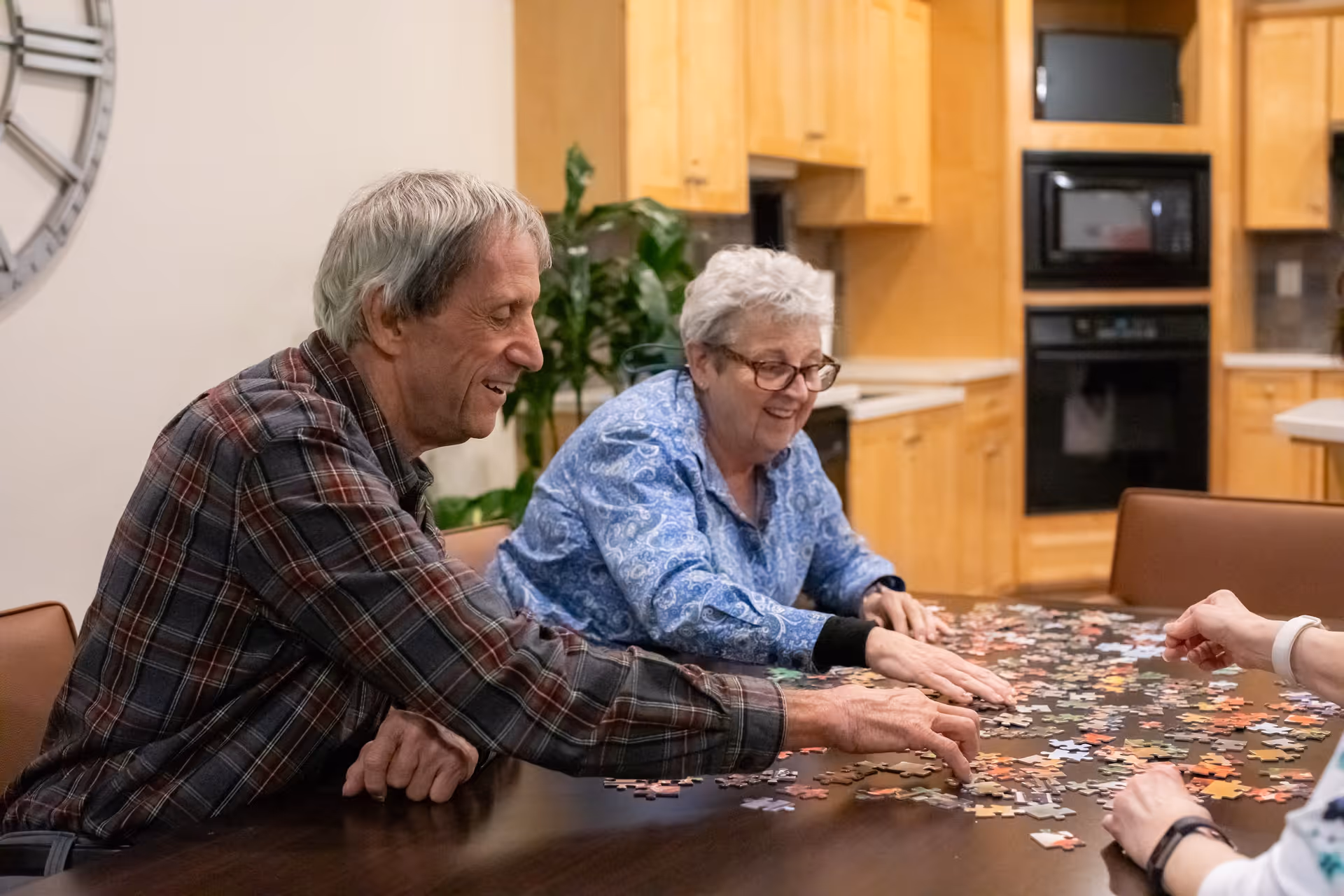 Two elderly individuals sitting at a table in a kitchen area, smiling and working together on assembling a jigsaw puzzle. The background shows wooden kitchen cabinets, a built-in oven, and a microwave.