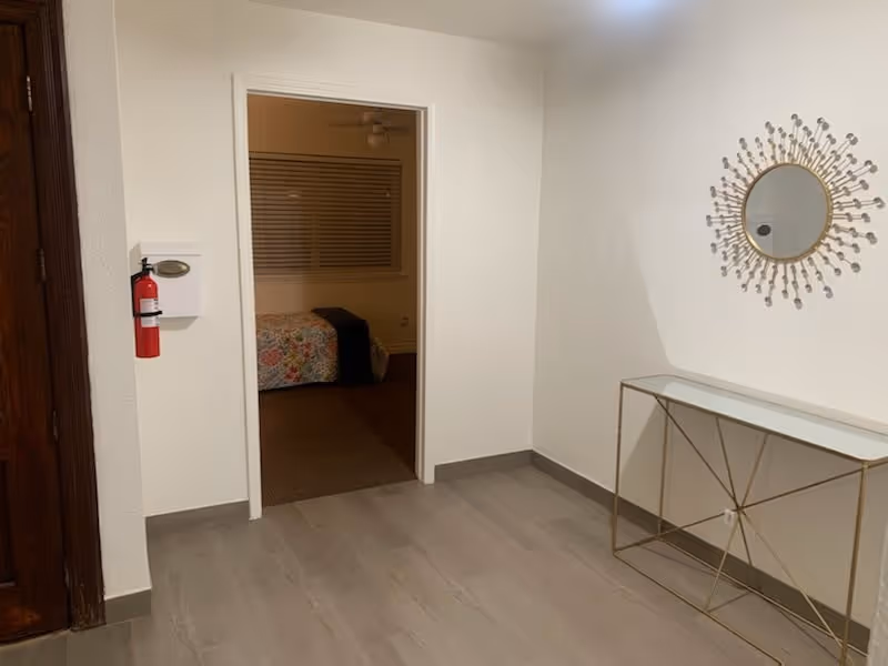 Entryway with gray wood-look floor, a decorative round mirror and console table, and an open doorway showing a bed in an adjacent bedroom.