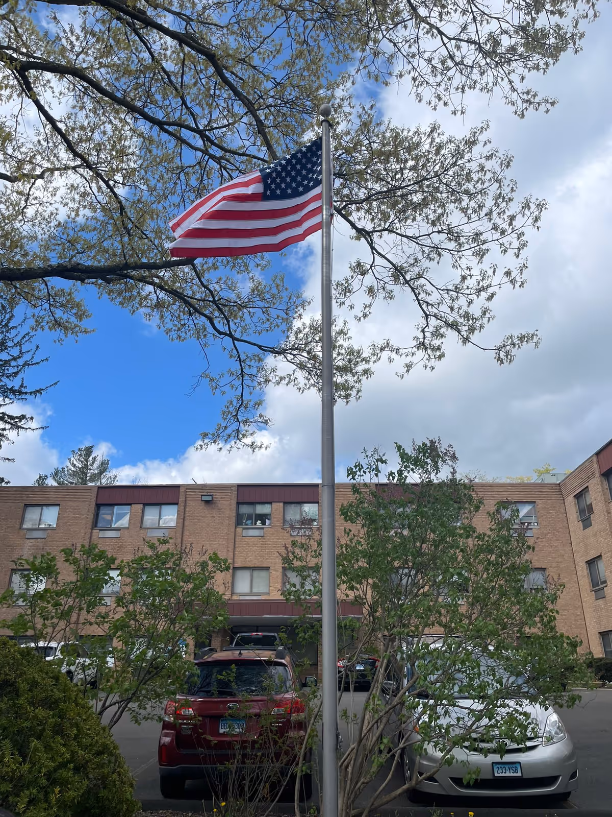 An American flag on a flagpole in front of a three-story brick building with several windows. There are trees and bushes around the flagpole, and two cars parked in front of the building under a partly cloudy sky.