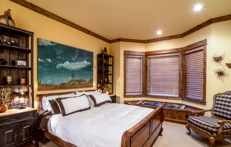 Warm bedroom featuring a wooden bed with white bedding, a bay window with wooden blinds and bench, wooden shelving and a plaid armchair.