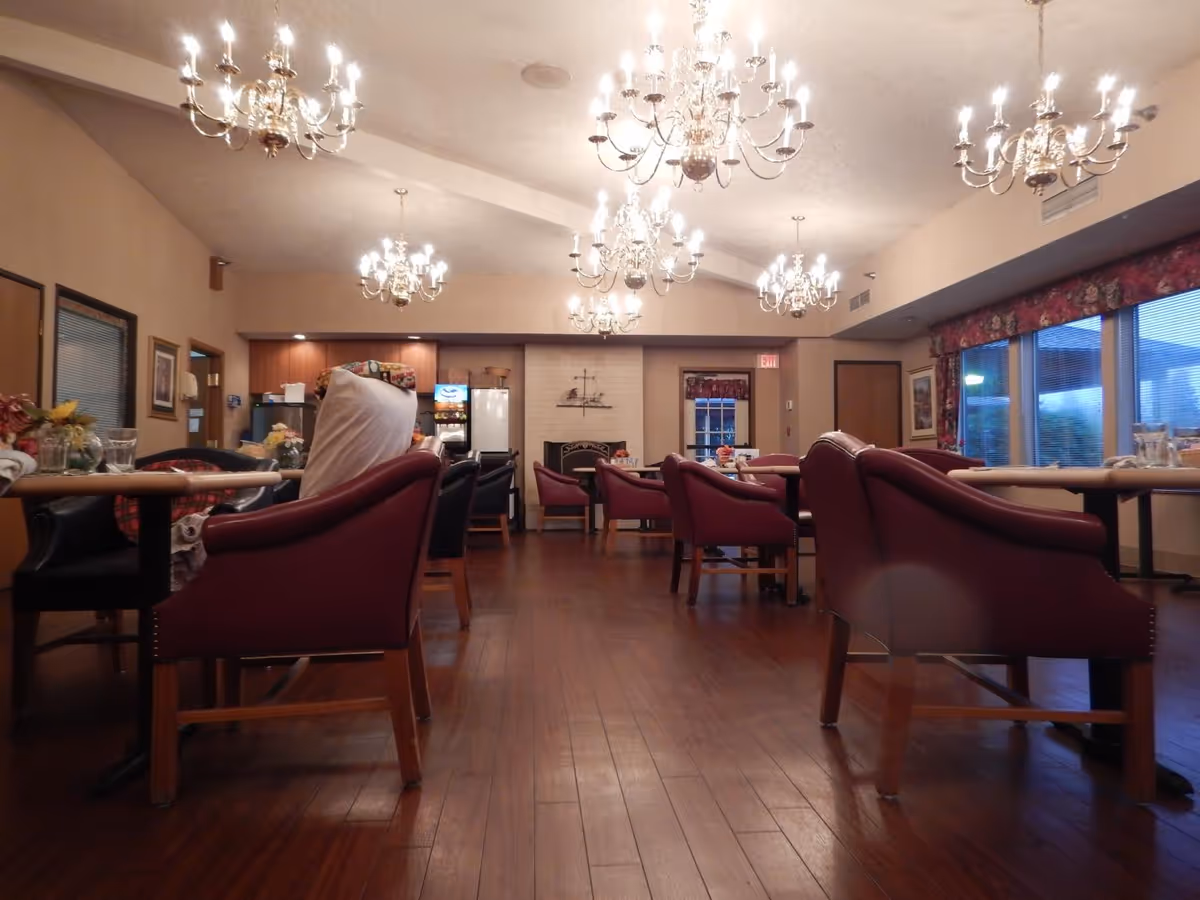Interior view of a dining room in an assisted living facility with multiple tables and chairs arranged neatly. The room features polished wooden floors, several ornate chandeliers hanging from the ceiling, large windows with floral curtains, and a fireplace at the far end. There are some decorative items and a beverage machine visible in the background.