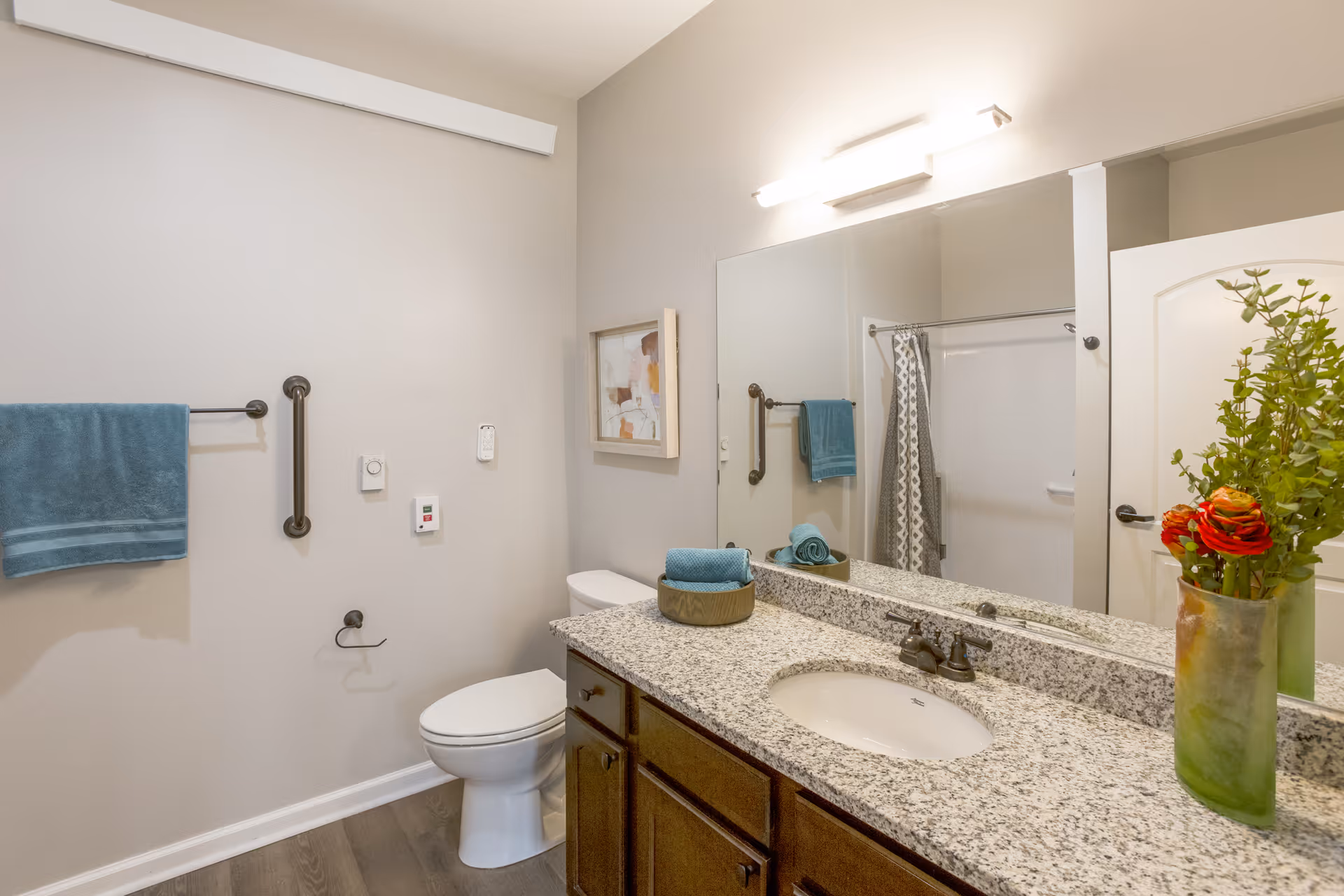 A clean and modern bathroom featuring a granite countertop with a built-in sink, a large mirror above the sink, a toilet, a towel rack with a blue towel, a grab bar, a shower with a patterned curtain, and a vase with red and orange flowers on the countertop.