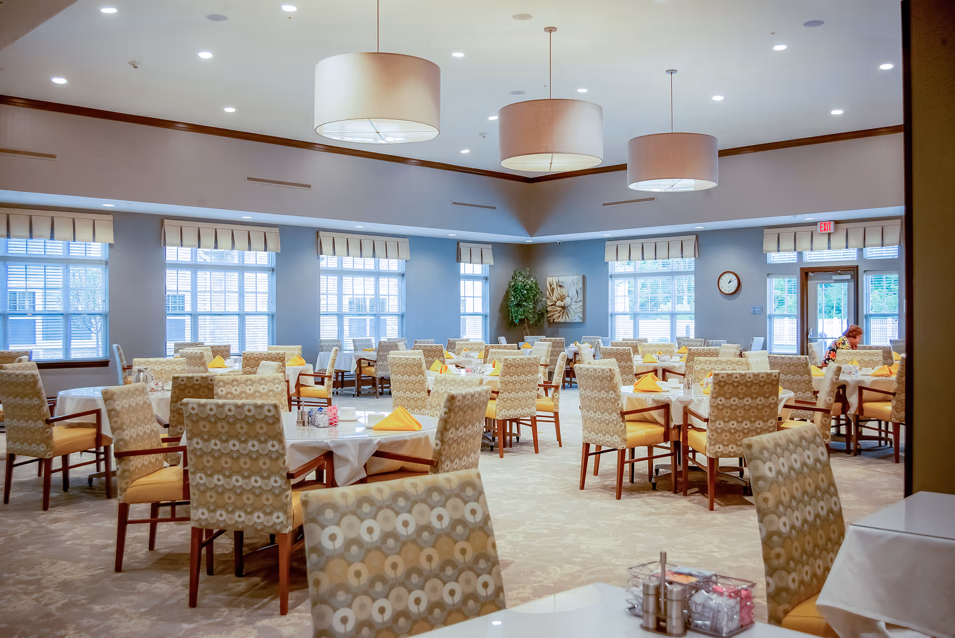 Spacious dining room with multiple round tables set with yellow napkins and patterned chairs beneath large pendant lights.
