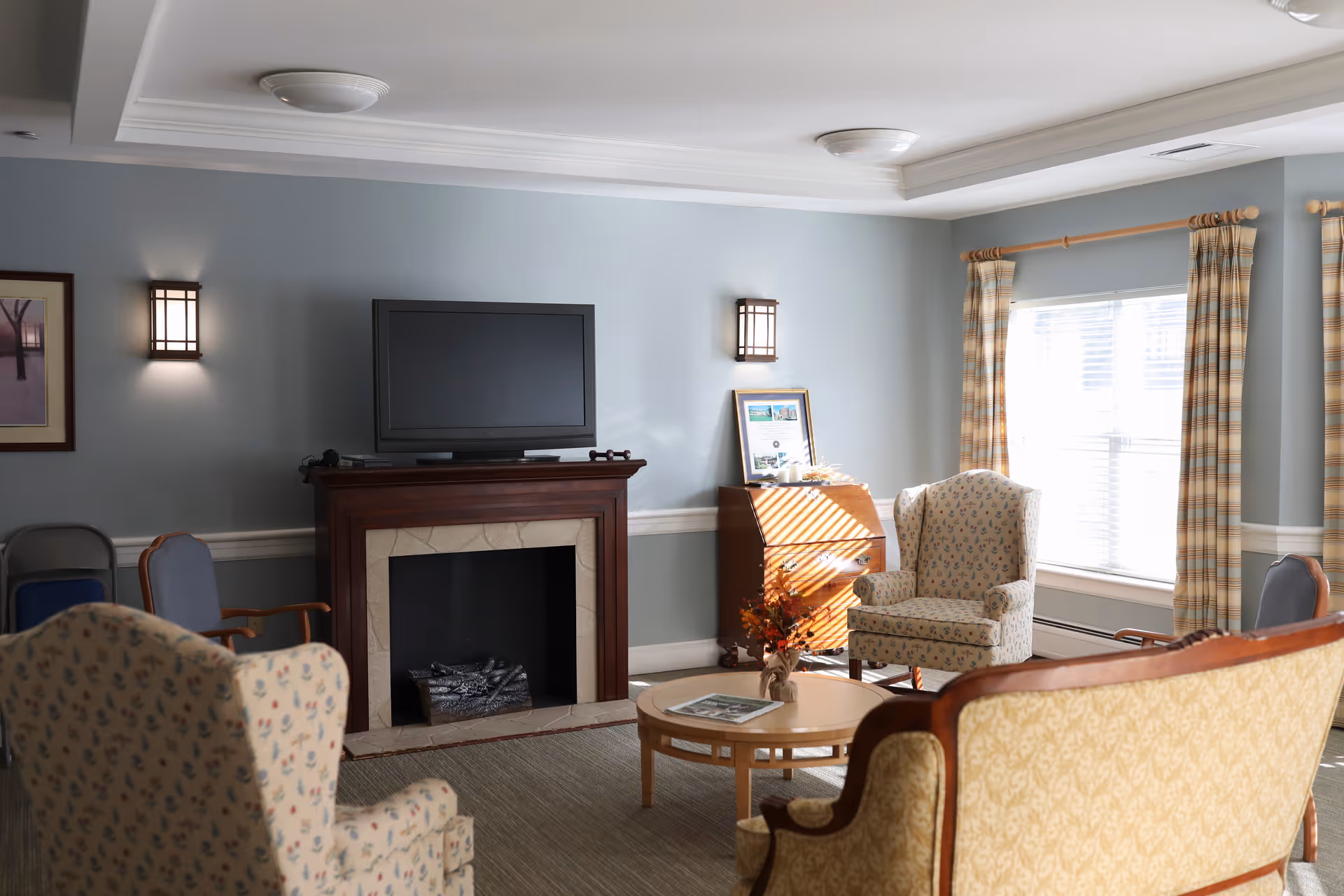 A cozy living room with light blue walls, a fireplace with a mounted flat-screen TV above it, two patterned armchairs, a wooden coffee table with a small flower arrangement, a wooden cabinet, and large windows with plaid curtains letting in natural light.