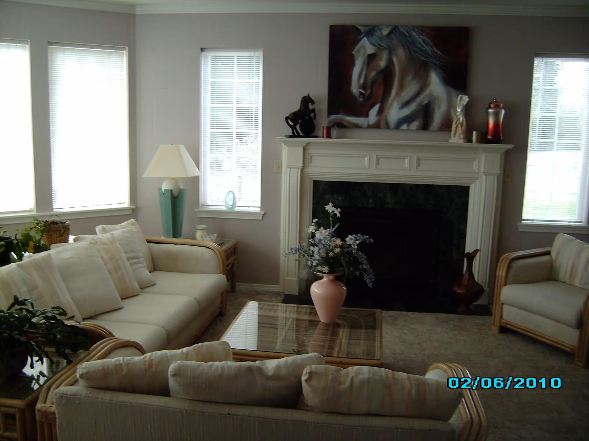 A cozy living room with beige cushioned sofas and armchairs arranged around a glass coffee table with a pink vase of flowers. The room features a white fireplace with a dark marble surround, decorated with a horse painting above it and various decorative items on the mantel. There are three tall windows with blinds letting in natural light, and a floor lamp next to one of the sofas.