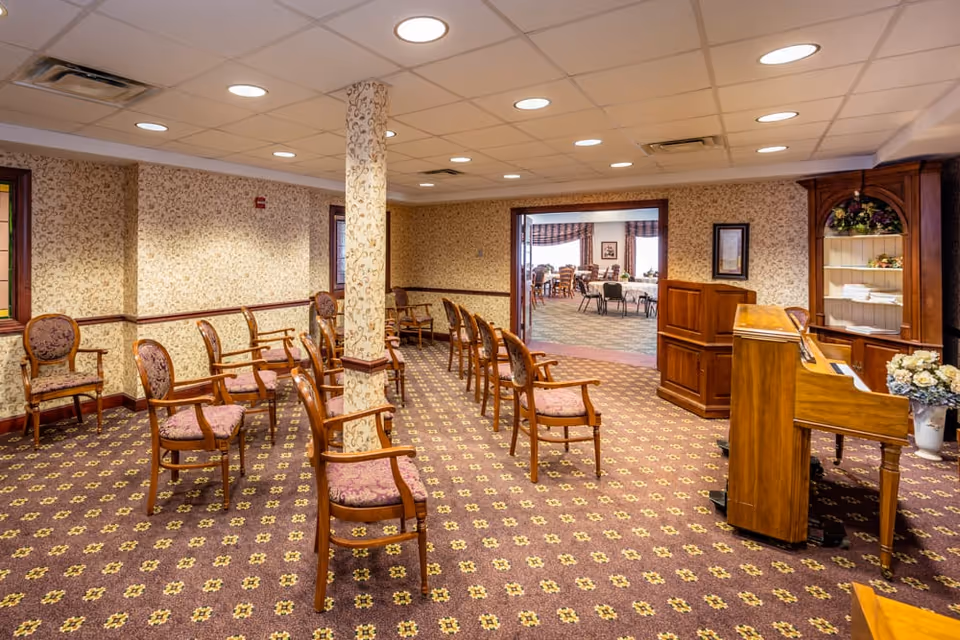 A room with multiple wooden chairs arranged in rows facing a wooden piano on the right side. The room has patterned wallpaper and carpet with floral designs. There is a pillar in the middle of the room and an open doorway leading to another room with tables and chairs.