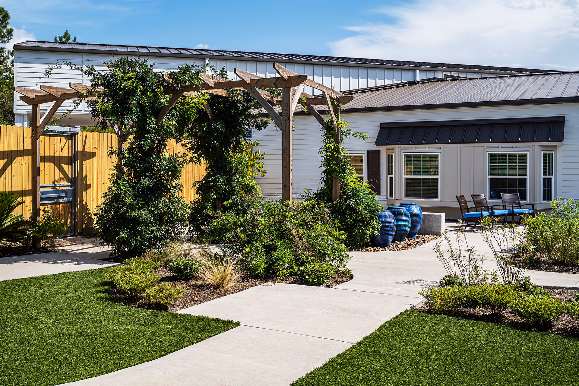 Outdoor garden area at Meredith Court featuring a wooden pergola with climbing plants, manicured green lawns, various shrubs and plants, a concrete pathway, and a building with white siding and multiple windows in the background. There are also blue ceramic pots and outdoor seating with chairs and cushions near the building.