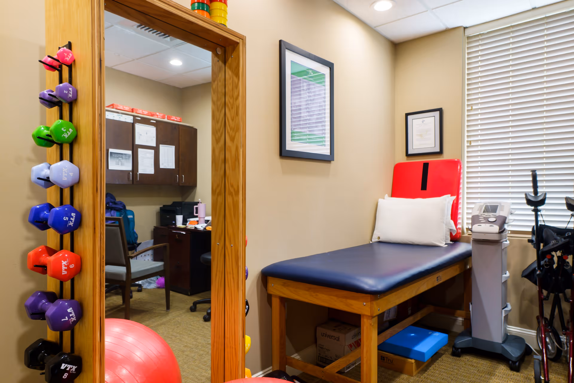 A small therapy or examination room with a padded treatment table featuring a red backrest and white pillow. To the left, a wooden rack holds various colorful dumbbells, and a large mirror reflects an adjacent office with a desk and chair. The room has beige walls, framed pictures, a window with blinds, and medical equipment including a machine and walkers.
