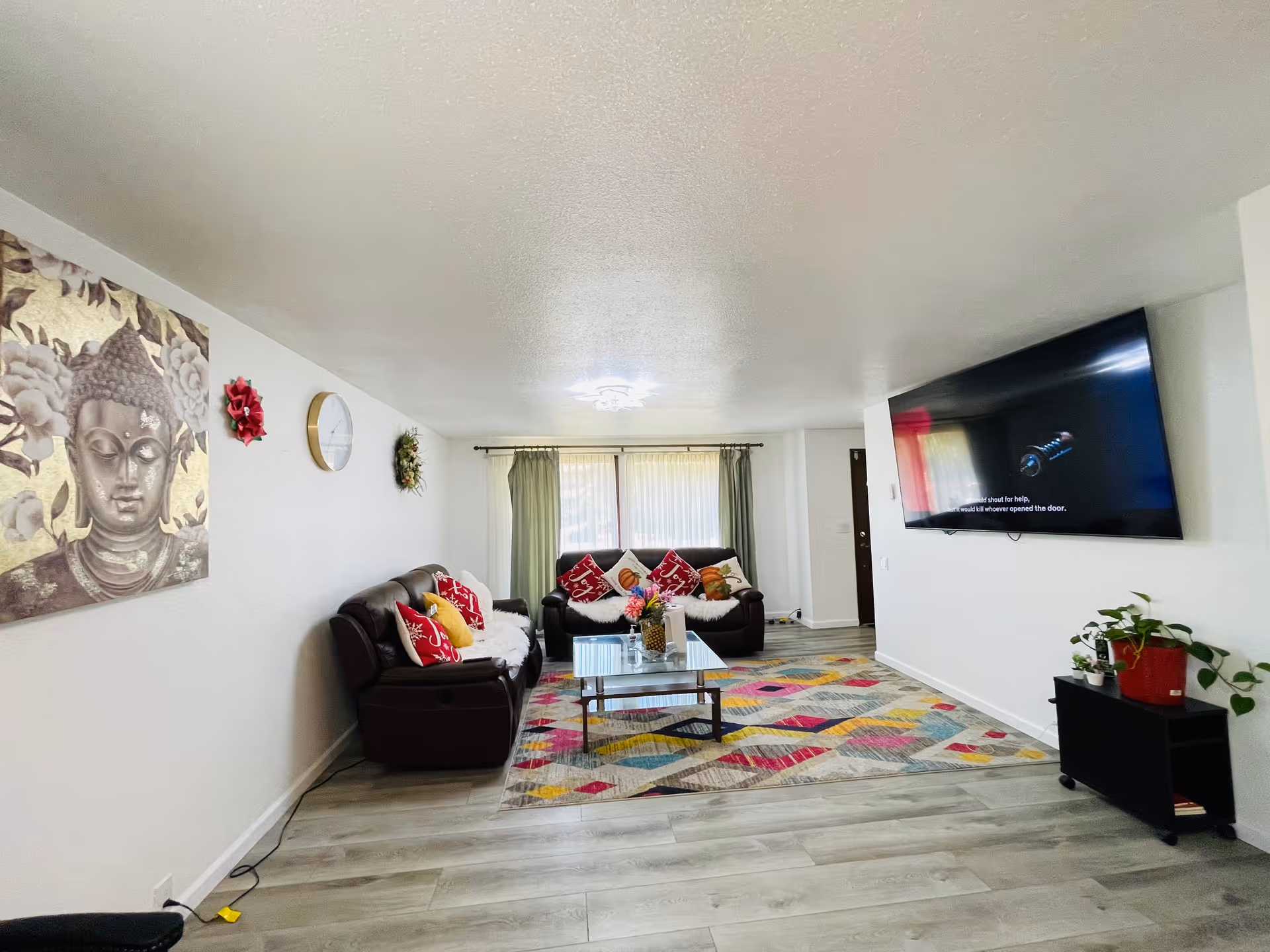 A bright living room with two dark brown leather sofas adorned with red and yellow pillows. A colorful geometric patterned rug covers the light wood floor. A glass coffee table with a flower arrangement sits in the center. On the left wall, there is a large Buddha painting, a clock, and decorative wreaths. A large flat-screen TV is mounted on the right wall above a small black stand with potted plants. Light filters in through a window with green curtains at the far end of the room.