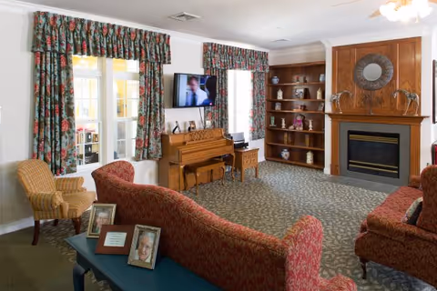 A cozy living room with patterned red sofas, a wooden piano, a flat-screen TV mounted on the wall, and a fireplace with a decorative mirror above it. There are two windows with floral curtains, a wooden bookshelf with various decorative items, and a small table with framed photos in the foreground.