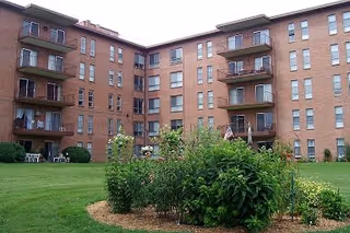 Exterior view of a multi-story brick apartment building with balconies overlooking a well-maintained grassy courtyard featuring a garden bed with various plants and flowers.